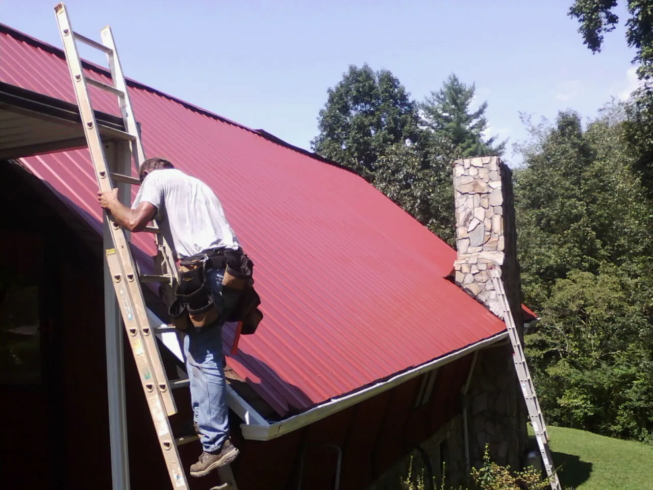Roofer on a ladder beside a red metal roof and stone chimney — Soffit & Fascia Repair by Roofing Brothers in Forest Ranch